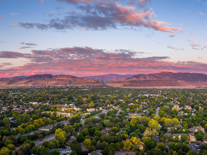 Suburban neighborhood with dense trees under a colorful sunset sky and distant mountains.
