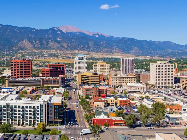 Downtown cityscape with mid-rise buildings and mountains in the background under a clear blue sky.