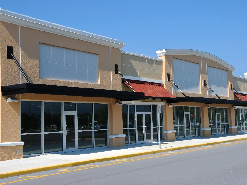 Empty storefronts in a beige commercial strip mall under a clear blue sky.